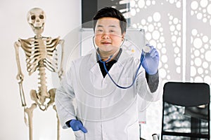 Asian male doctor in white coat, protective gloves and stethoscope in his hand, standing in his office with big human