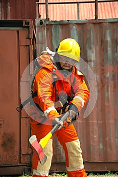 An Asian male fireman in orange protective clothing, mask and helmet with an ax standing front of building.
