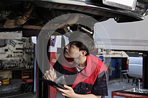 Asian male motor mechanic inspects undercarriage of EV car at service garage.