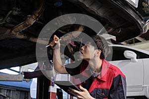 Asian male motor mechanic inspects undercarriage of EV car at service garage.
