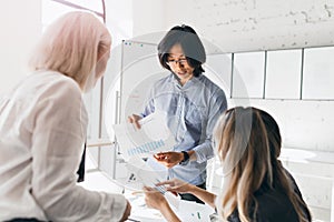 Asian male office worker with wristwatch holding documents with diagrams while talking with female colleagues. Indoor