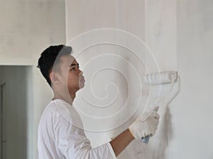 Asian male painter painting new white wall with roller brush, house interior renovation