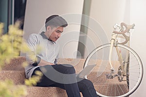 Asian male student sitting at the stairs and smiling as use laptop computer with his bike background at campus,Students and