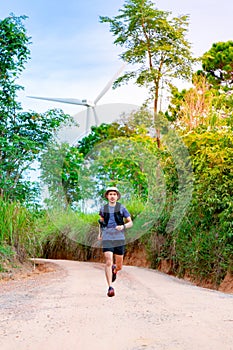 An Asian male trail runner practicing on a dirt road in a forest behind trees and windmills generating electricity