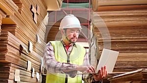 Asian male worker in uniform inspects with laptop at paper factory warehouse.