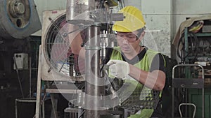 An Asian male worker using a drilling machine in a factory. He wears glasses and a safety helmet for working. engineering and indu