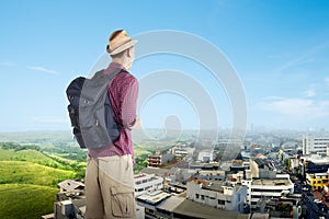 Asian man with a backpack looking at green landscapes and a city with a blue sky background