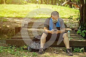Asian man in black casual wear wearing a face mask sitting on old boards of a wooden bridge using an application on cell phone in