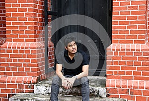 Asian man in black shirt sitting in front of a black door between two brick pillars.