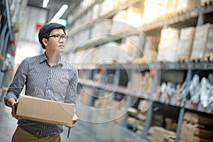 Asian man carrying cardboard box between row of shelves in warehouse, shopping warehousing or working pick and packing concepts