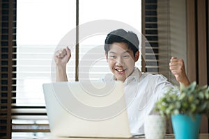 Asian man celebrating success while working on laptop at home.
