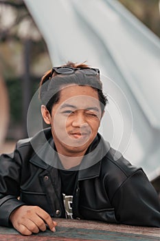 an Asian man with a chubby face wearing sunglasses and a black leather jacket while sitting at a cafe table