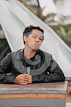 an Asian man with a chubby face wearing sunglasses and a black leather jacket while sitting at a cafe table