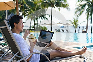 Asian man drinking coconut water while he is working on his laptop on a chairnear pool at resort hotel on summer vacation holiday