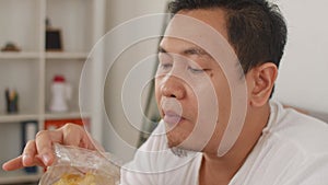 Asian man enjoys eating casava or potato chips while sitting on sofa at living room