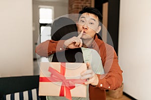 Asian man got surprise for his loving wife, husband embracing woman, holding gift box and showing silence gesture