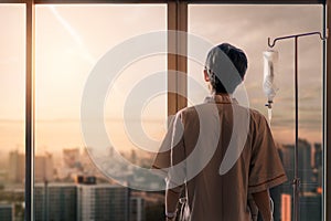 Asian man hand of a patient standing with medical drip or IV drip in hospital ward and look out at window with sunrise time,