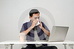 asian man has a runny nose, wipes his nose with tissue paper at workplace while sitting in front of laptop computer. 