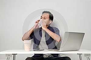 asian man has a runny nose, wipes his nose with tissue paper at workplace while sitting in front of laptop computer. 