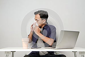 asian man has a runny nose, wipes his nose with tissue paper at workplace while sitting in front of laptop computer. isolated
