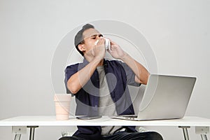asian man has a runny nose, wipes his nose with tissue paper at workplace while sitting in front of laptop computer. isolated