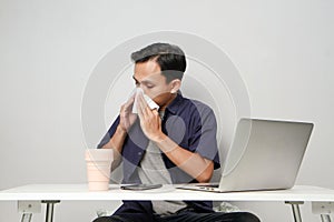 asian man has a runny nose, wipes his nose with tissue paper at workplace while sitting in front of laptop computer. isolated