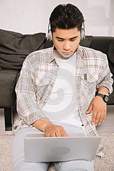 Asian man in headphone using laptop while sitting on floor at home