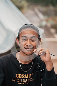 an Asian man with his hair tied up and wearing a black sweater jacket is sitting relaxing at a cafe table when meeting his friends