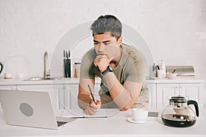 Asian man holiding pencil while looking at laptop in kitchen