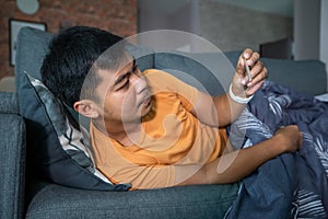 Asian man is ill and coughs on the sofa at home and he uses a thermometer to check the temperature during the coronavirus epidemic