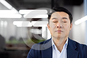 Asian man inside office close up, mature boss in business suit with closed eyes meditating, thinking and solving