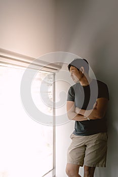 Asian man leaning against white wall in navy blue t-shirt and khaki shorts, looking out of the windows.