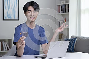 Asian man looking at camera while holding credit card and using laptop computer.