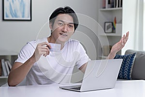 Asian man looking at camera while holding credit card and using laptop computer. Businessman working at home. Online shopping