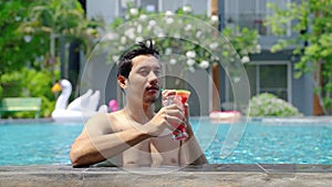 Asian man lounging in the hotel pool happily sipping juice on his vacation