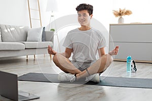 Asian man meditating in lotus position on mat with pc