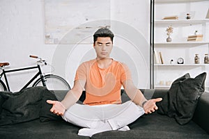 Asian man meditating while sitting in lotus pose on sofa at home