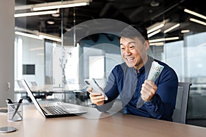 An Asian man in office holding cash money and phone. Rejoicing at the win, placing bets online