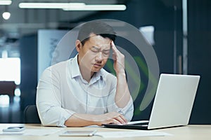An asian man office worker, working on a laptop. He holds his head, feels a headache