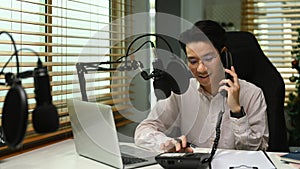 Asian man radio host talking on telephone and using condenser microphone recording voice conversation at home studio