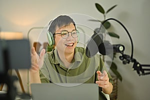 Asian man radio host using laptop and talking through microphone to recording podcast in small home studio