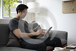 Asian man resting on couch and browsing internet with computer tablet.
