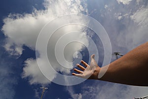 an Asian man's hand is trying to reach the clear blue sky with beautiful white clouds in the midday.