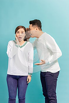 Asian man sharing secret or whispering gossips into his girlfriend`s ear, blue studio background