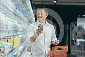 asian man shopper walks around the supermarket with a shopping cart looking at smart phone browsing smartphone in hands