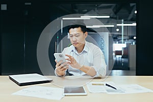 An Asian man sits in the office, holds the phone, dials a message, is upset and reads bad news.
