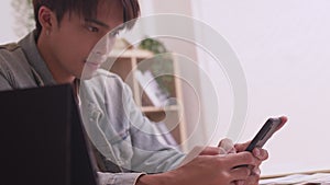 Asian man sits on sofa at home, texting and talking with buddy, holding smartphone,or checking mobile app or social media