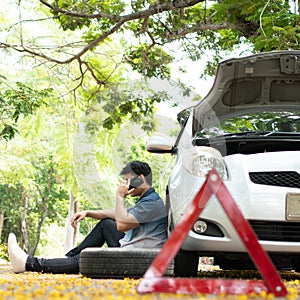 Asian man sitting beside car and using mobile phone calling for assistance after a car breakdown on street. Concept of vehicle