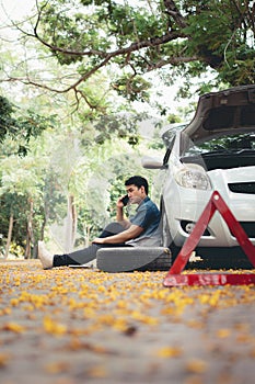Asian man sitting beside car and using mobile phone calling for assistance after a car breakdown on street. Concept of vehicle