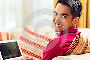 Asian man sitting on couch surfing the internet